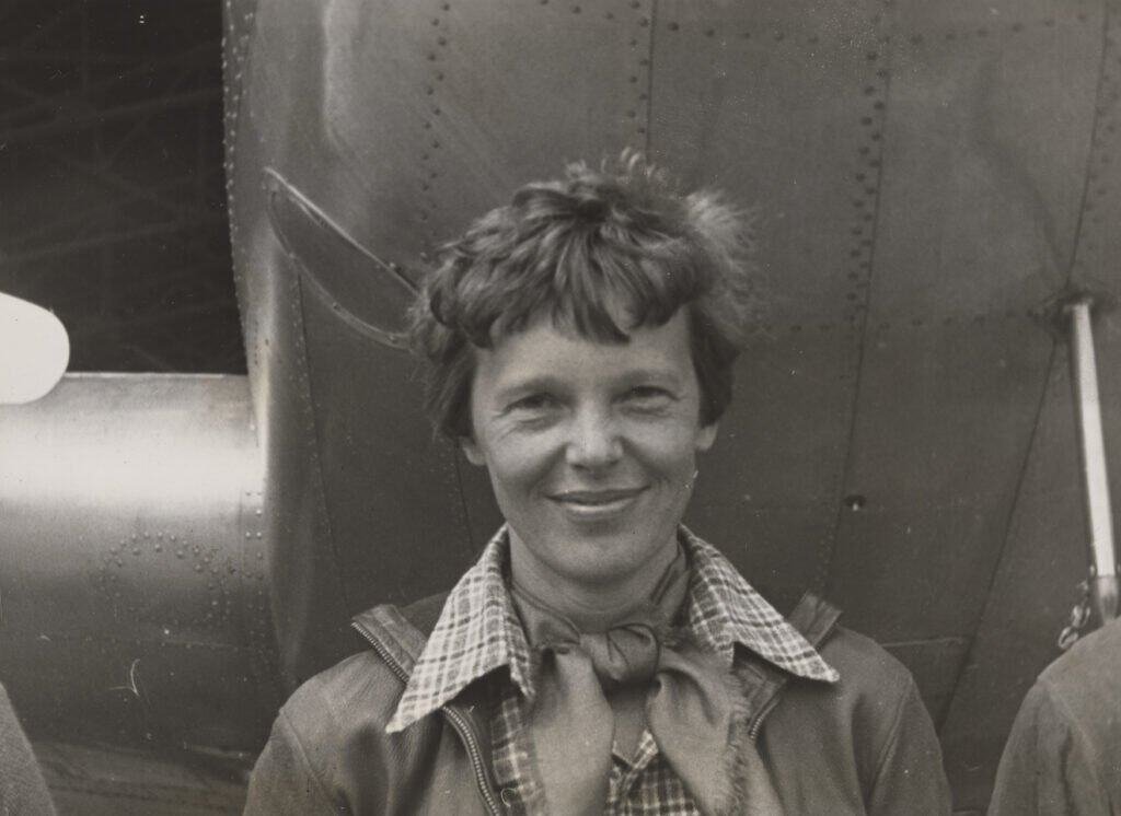 Amelia Earhart em pé sob o nariz do seu avião Lockheed Model 10-E Electra